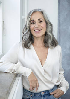 Smiling woman with gray hair standing by window - JBYF00139