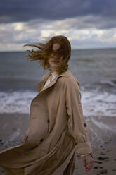 Frau mit zerzaustem Haar und Trenchcoat am Strand - SSGF00559