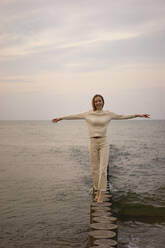 Happy woman with arms outstretched walking on wooden post at beach - SSGF00575