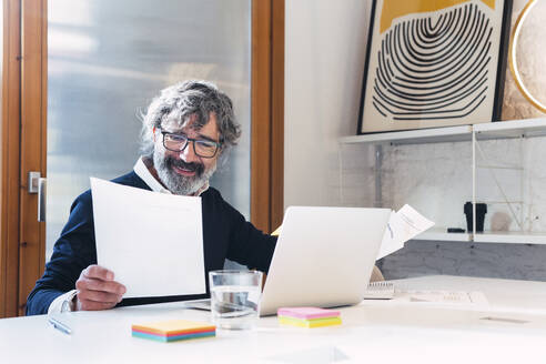 Smiling freelancer reading documents sitting with laptop at table - PNAF03525