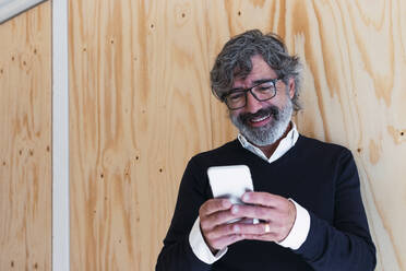 Lächelnder älterer Mann, der mit seinem Smartphone vor einer Wand Textnachrichten verschickt - PNAF03550