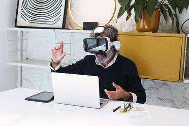 Freelancer wearing virtual reality simulator gesturing sitting with laptop at home - PNAF03558