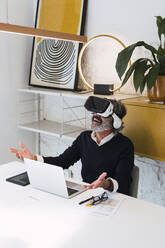Smiling freelancer wearing virtual reality simulator gesturing sitting with laptop at table - PNAF03559