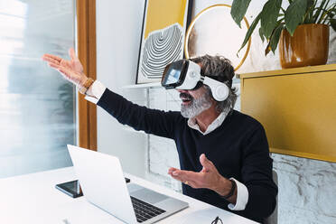 Glücklicher älterer Freiberufler, der einen Virtual-Reality-Simulator trägt und gestikulierend mit einem Laptop am Tisch sitzt - PNAF03560