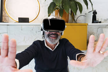 Happy senior freelancer wearing virtual reality simulator gesturing at home - PNAF03561