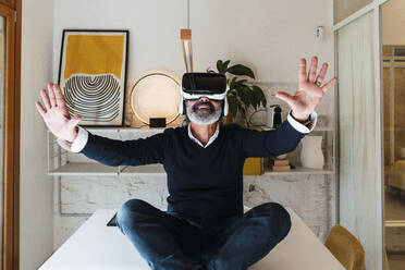 Glücklicher Freiberufler, der einen Virtual-Reality-Simulator trägt und gestikulierend auf einem Tisch zu Hause sitzt - PNAF03562