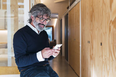 Glücklicher älterer Mann, der eine Textnachricht über sein Smartphone verschickt und zu Hause sitzt - PNAF03567
