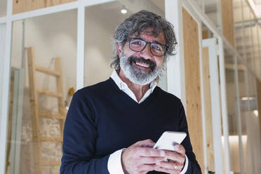 Smiling senior man wearing eyeglasses holding mobile phone at home - PNAF03570