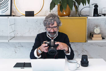 Senior freelancer holding camera sitting with laptop at home - PNAF03583
