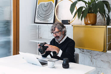 Lächelnder Freiberufler mit Kamera in der Hand und Laptop am Tisch - PNAF03584