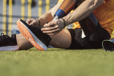 Hände eines Sportlers, der die Schnürsenkel einer Beinprothese im Gras sitzend bindet - JCCMF05984