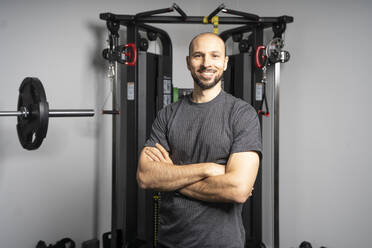 Lächelnder Fitnesslehrer, der mit verschränkten Armen vor einem Trainingsgerät im Fitnessstudio steht - DAMF00913