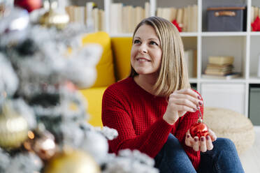 Smiling woman with Christmas bauble sitting in living room at home - GIOF15205