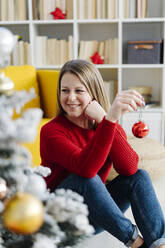 Smiling woman with hand on chin holding Christmas bauble in living room at home - GIOF15206