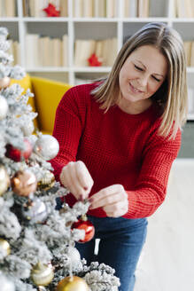 Lächelnde Frau schmückt Weihnachtsbaum mit Christbaumkugel zu Hause - GIOF15207