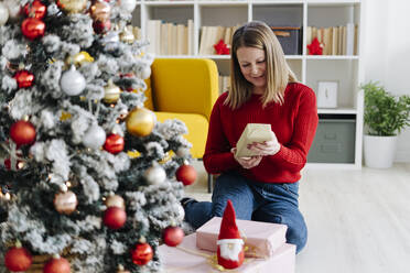 Smiling woman with gifts sitting by Christmas tree at home - GIOF15210