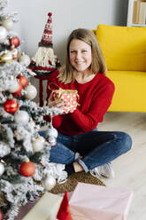 Lächelnde Frau mit Weihnachtsgeschenk unter dem geschmückten Baum zu Hause - GIOF15213