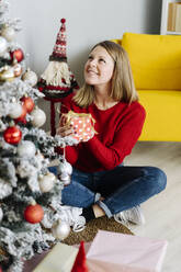 Lächelnde Frau mit Weihnachtsgeschenk und Blick auf den geschmückten Baum im heimischen Wohnzimmer - GIOF15214