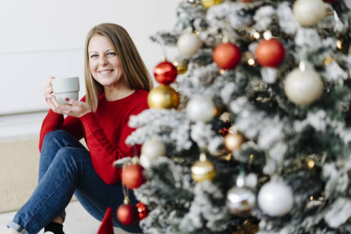 Glückliche Frau mit Kaffeetasse unter dem Weihnachtsbaum zu Hause - GIOF15217