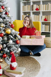 Lächelnde Frau mit Hand am Kinn sitzt mit Geschenken im Wohnzimmer zu Hause - GIOF15224