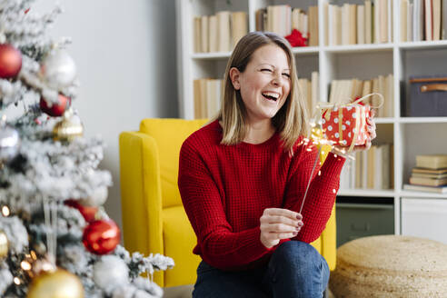 Fröhliche Frau mit Wunderkerze und Weihnachtsgeschenk im heimischen Wohnzimmer sitzend - GIOF15228