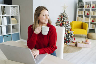 Lächelnde Frau mit Kaffeetasse und Laptop am Wohnzimmertisch sitzend - GIOF15230