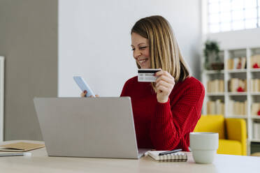 Happy woman using mobile phone holding credit card at table in living room - GIOF15239