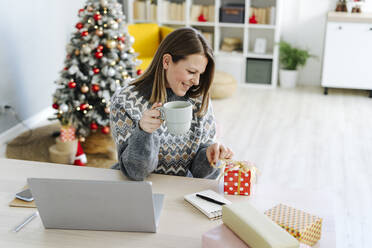 Lächelnde Frau hält eine Kaffeetasse und betrachtet ein Weihnachtsgeschenk - GIOF15243