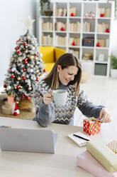 Lächelnde Frau betrachtet das Weihnachtsgeschenk auf dem Tisch im Wohnzimmer - GIOF15244