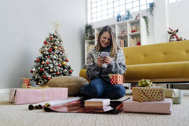 Happy woman using mobile phone sitting by gifts at home - GIOF15251