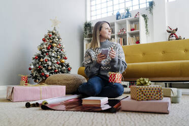 Woman with mobile phone sitting by gifts in living room at home - GIOF15252