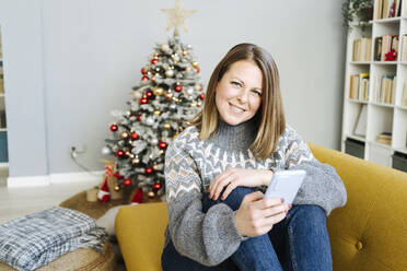 Glückliche blonde Frau mit Handy sitzt auf dem Sofa im Wohnzimmer zu Hause - GIOF15265