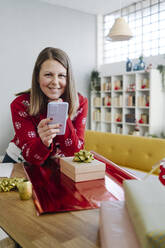 Lächelnde Frau mit Handy auf dem Tisch im Wohnzimmer zu Hause gelehnt - GIOF15271