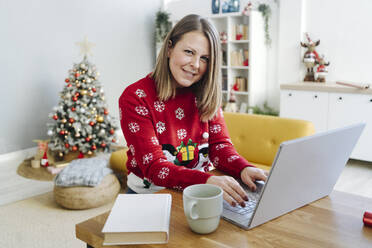 Lächelnde blonde Frau mit Laptop am Tisch im Wohnzimmer sitzend - GIOF15273
