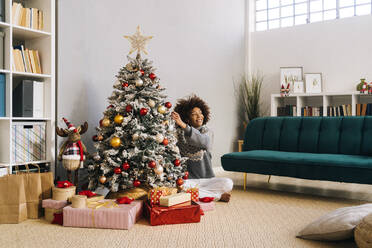 Lächelnde junge Frau im Schneidersitz beim Schmücken des Weihnachtsbaums zu Hause im Wohnzimmer - GIOF15282