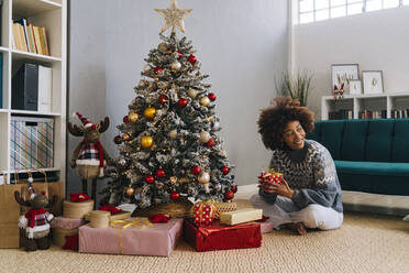 Happy woman sitting with gifts by Christmas tree at home - GIOF15290