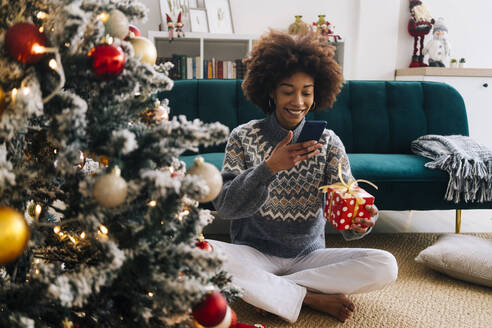 Smiling young woman photographing Christmas present through mobile phone at home - GIOF15291