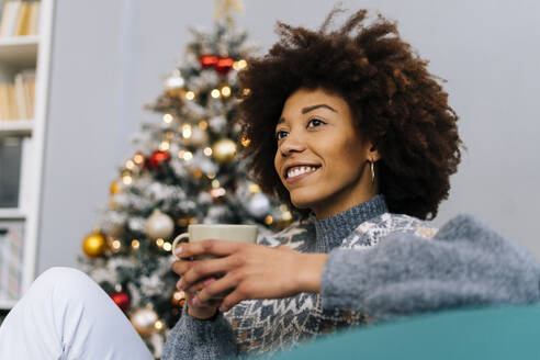 Lächelnde junge Frau mit Kaffeetasse, die zu Hause auf dem Sofa sitzt - GIOF15300