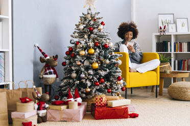 Young woman with note pad sitting on yellow armchair in living room at home - GIOF15302