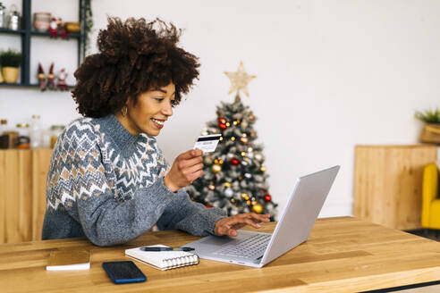 Happy young woman with credit card using laptop sitting at table in living room - GIOF15322