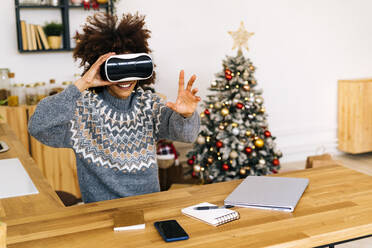 Smiling woman wearing virtual reality simulator gesturing sitting at table in living room - GIOF15327