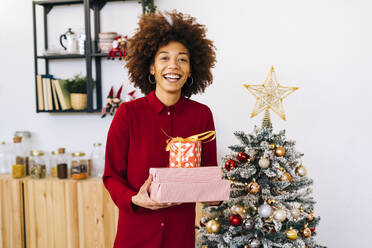 Happy woman with gifts standing by Christmas tree at home - GIOF15341