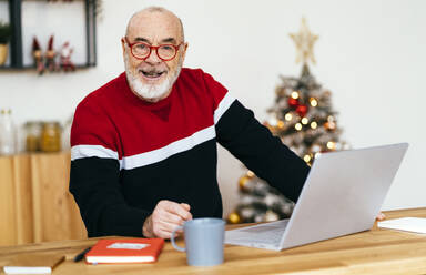 Smiling senior man with laptop sitting at home - GIOF15356