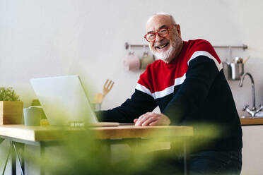 Glücklicher älterer Mann mit Brille sitzt mit Laptop zu Hause - GIOF15357