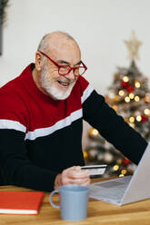 Happy senior man holding credit card doing online shopping through laptop at home - GIOF15358