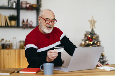 Senior man holding credit card doing online shopping through laptop at home - GIOF15359