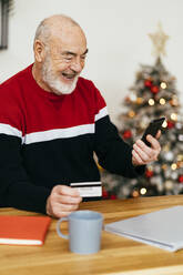 Glücklicher älterer Mann mit Kreditkarte beim Online-Shopping mit dem Handy zu Hause - GIOF15362