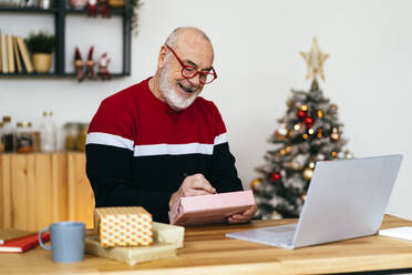 Happy senior man writing on gift box sitting with laptop at table at home - GIOF15364