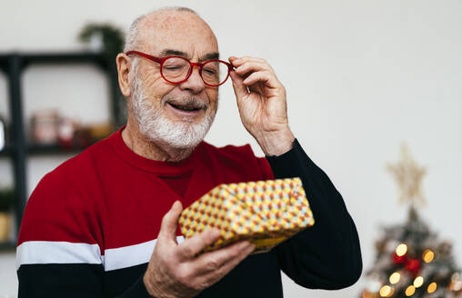 Lächelnder älterer Mann mit Brille betrachtet eine Geschenkbox zu Hause - GIOF15365