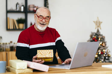 Senior man holding gift box using laptop at home - GIOF15366